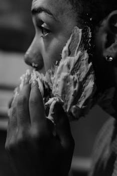 Artistic black and white photo of a man applying shaving foam to his face, emphasizing texture and detail.